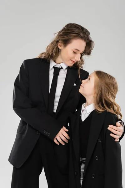 Crianza moderna, mujer de negocios alegre en traje abrazando a la hija en uniforme escolar y de pie juntos sobre un fondo gris, madre e hijo felices, de vuelta a la escuela - foto de stock