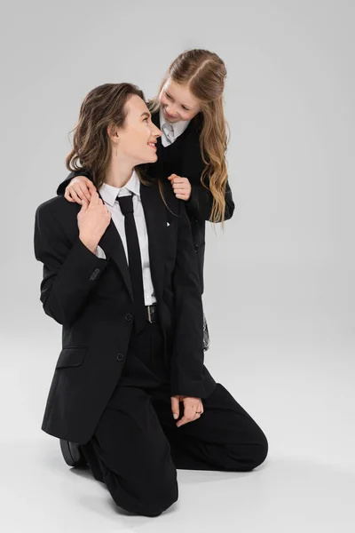Happy schoolgirl hugging working mother, cheerful girl in school uniform looking at mom in suit on grey background, formal attire, fashionable family, bonding, modern parenting — Stock Photo