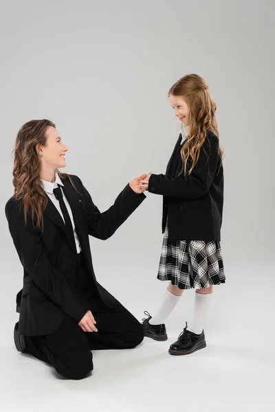 Smiling schoolgirl holding hands with working mother, cheerful girl in school uniform looking at mom in suit on grey background, formal attire, fashionable family, bonding, modern parenting — Stock Photo