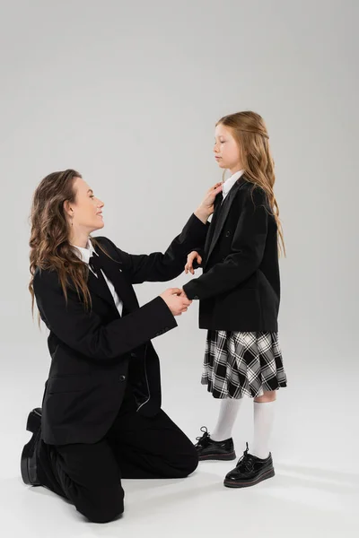 Mother adjusting tie of schoolgirl, happy woman in business attire and daughter on grey background in studio, back to school, getting ready, school preparation, modern parenting, parent-child — Stock Photo