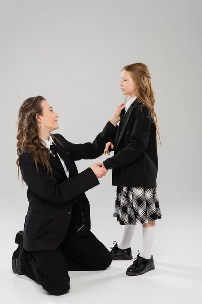 Madre che aggiusta il legame di scolaretta, donna felice in abito da lavoro e figlia su sfondo grigio in studio, torna a scuola, si prepara, preparazione scolastica, genitorialità moderna, genitore-bambino — Foto stock