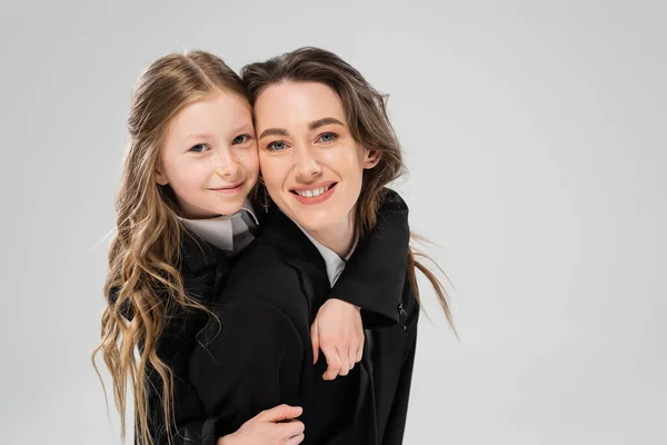 Cheerful schoolgirl hugging mother in suit, girl in school uniform and her mom in business attire on grey background, fashionable family, bonding, modern parenting, back to school, portrait — Stock Photo