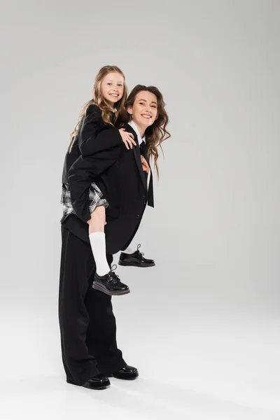 Cheerful mother piggybacking her daughter, working mom in business attire and schoolgirl in uniform on grey background in studio, modern parenting, fashionable family, having fun — Stock Photo