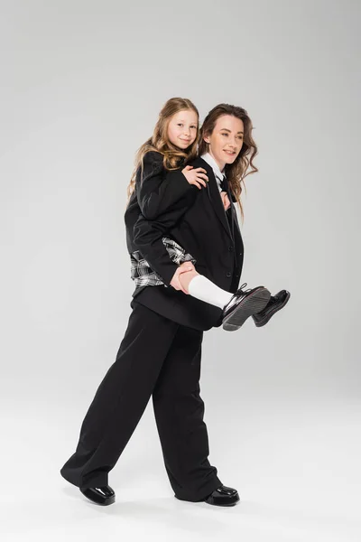 Mother piggybacking daughter in school uniform, working mom in business attire and schoolgirl on grey background in studio, modern parenting, fashionable family, having fun — Stock Photo