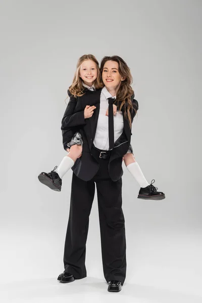Mãe trabalhando piggybacking sua filha feliz, empresária em traje formal e estudante em uniforme em fundo cinza no estúdio, parentalidade moderna, família na moda, se divertindo — Fotografia de Stock