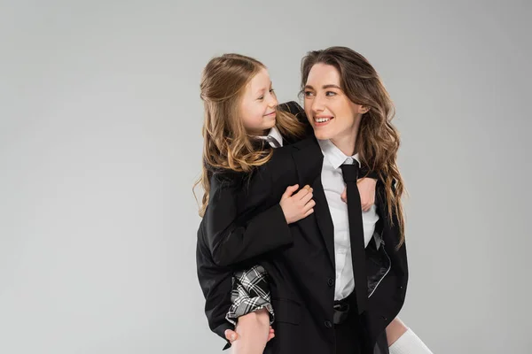 Crianza moderna, madre feliz apoyando a su hija, mamá trabajadora en traje de negocios y colegiala en uniforme sobre fondo gris en el estudio, familia de moda, divertirse - foto de stock
