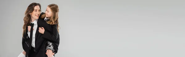 Madre sorridente a cavalluccio sua figlia, mamma che lavora in abiti da lavoro e studentessa in uniforme su sfondo grigio in studio, genitorialità moderna, famiglia alla moda, divertirsi, banner — Foto stock