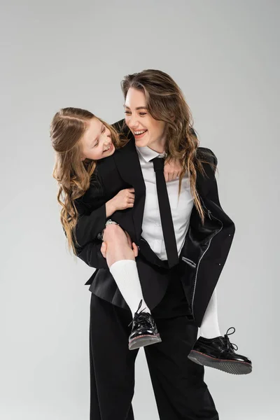 Mãe e filha se divertindo, mulher alegre piggybacking estudante, trabalhando mãe em traje formal e criança em uniforme no fundo cinza no estúdio, parentalidade moderna — Fotografia de Stock