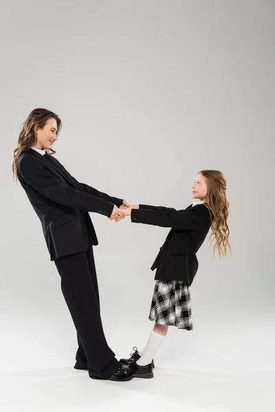 Mother and schoolgirl holding hands, happy woman in business attire and child in school uniform standing together on grey background, modern parenting, back to school — Stock Photo