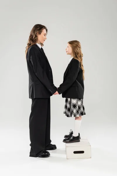 Мати і дитина тримаються за руки, щаслива жінка в діловому одязі і дитина в шкільній формі стоїть на стільці на сірому фоні, сучасне батьківство, обличчя до лиця — стокове фото