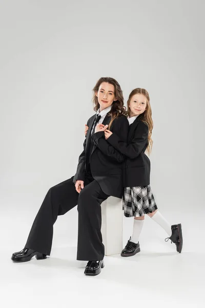 Glückliches Mädchen umarmt arbeitende Mutter, Geschäftsfrau sitzt auf Betonhocker neben Kind in Uniform auf grauem Hintergrund im Atelier, formelle Kleidung, Eltern-Kind, moderne Elternschaft, Modeshooting — Stockfoto