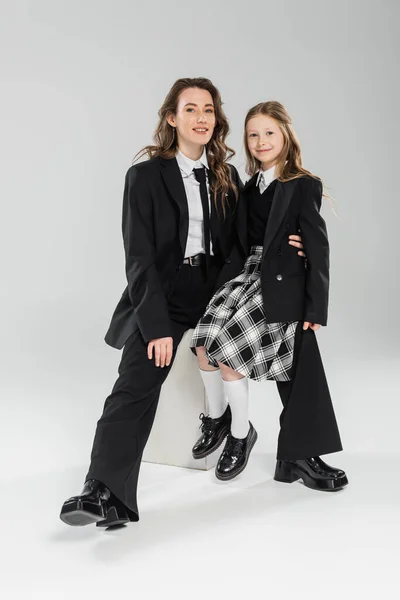 Working mother and daughter, happy businesswoman and schoolgirl sitting on concrete stool together on grey background in studio, formal attire, modern parenting, fashion shoot — Stock Photo