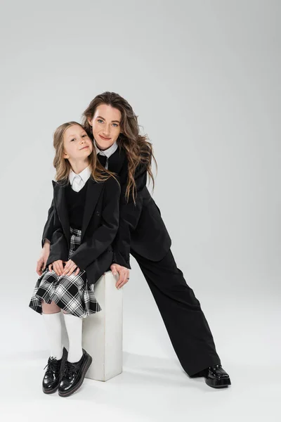 Mother and child, happy businesswoman in formal attire standing near schoolgirl sitting on concrete stool on grey background in studio, modern parenting, fashion shoot — Stock Photo