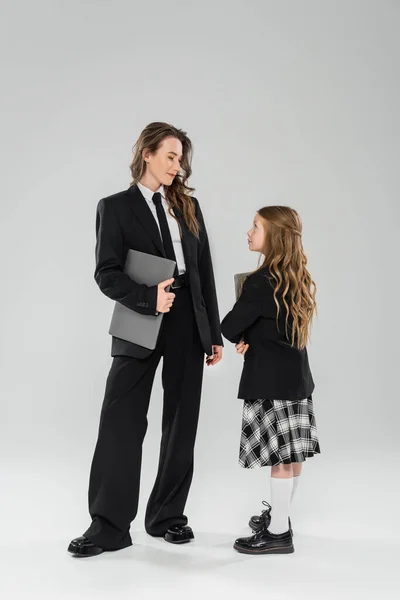 Working mother and daughter, digital nomadism, remote work, e learning, businesswoman in suit and schoolgirl standing together with laptops on grey background in studio, modern parenting — Stock Photo