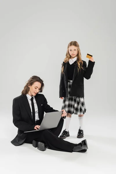 Working mother and daughter, digital nomadism, schoolgirl holding credit card near mom using laptop on grey background, modern parenting, business attire, financial education, online purchase — Stock Photo