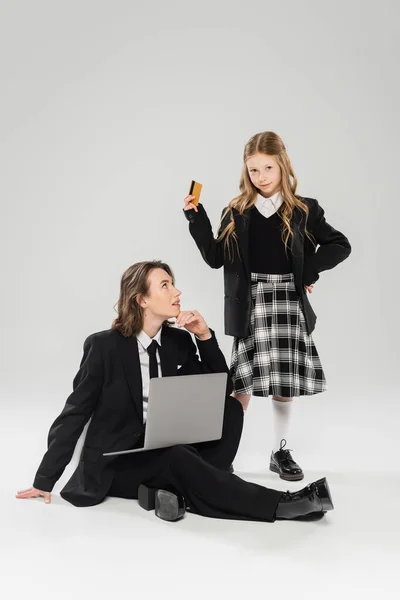 Mother and schoolgirl, digital nomadism, girl holding credit card near working mom using laptop on grey background in studio, modern parenting, business attire, online purchase, e-commerce — Stock Photo