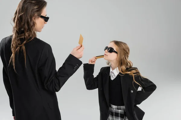 Mother and daughter in sunglasses, businesswoman in suit and schoolgirl in uniform holding credit cards and looking at each other on grey background, modern parenting, money management — Stock Photo