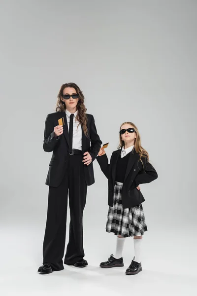 Budgeting, money management, mother and daughter in sunglasses, businesswoman in suit and schoolgirl in uniform holding credit cards on grey background, modern parenting, financial learning — Stock Photo