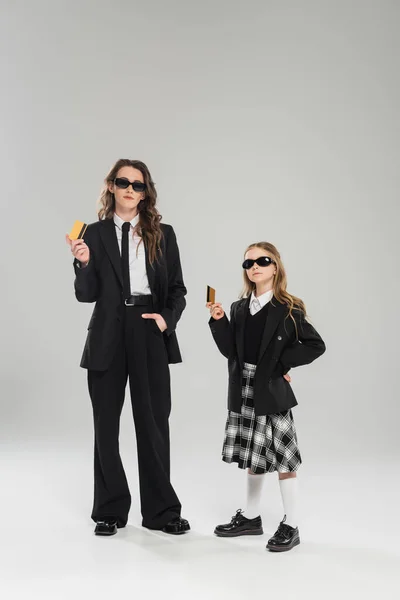 Mother and daughter in sunglasses, businesswoman in suit and schoolgirl in uniform holding credit cards on grey background, modern parenting, financial learning, budgeting, money management — Stock Photo