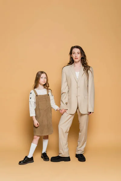 Mãe e filha na moda, mãe trabalhadora elegante em terno de mãos dadas com a filha enquanto estão juntos em fundo bege, pai e filho, família na moda — Fotografia de Stock