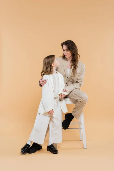 Madre e hija con estilo en trajes, mujer sentada en la silla alta y mirando a la chica en fondo beige, trajes de moda, atuendo formal, mamá corporativa, familia moderna — Stock Photo