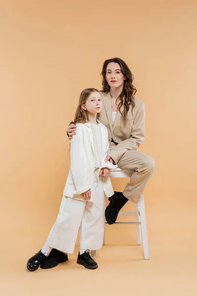 Madre e hija con estilo en trajes, mujer y niña mirando a la cámara, sentado en la silla alta sobre fondo beige, trajes de moda, atuendo formal, mamá corporativa, familia moderna - foto de stock