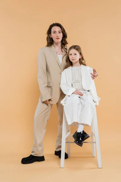 Élégante mère et fille en costumes, regardant la caméra tout en étant assis sur une chaise haute près de la femme sur fond beige, tenues à la mode, tenue formelle, maman d'entreprise, famille moderne — Photo de stock