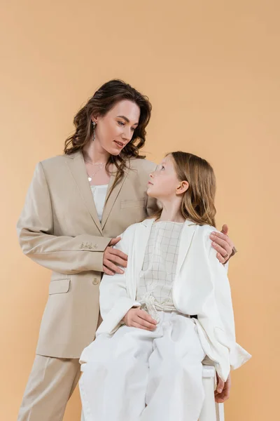 Business mother and daughter in suits, woman hugging shoulders of girl sitting on chair on beige background, fashionable outfits, formal attire, corporate mom, modern family — Stock Photo