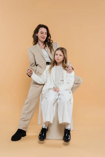 Stylish mother and daughter in suits, woman and girl holding hands and sitting together on beige background, fashionable outfits, formal attire, corporate mom, modern family — Stock Photo