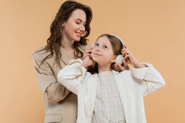Corporate mom and daughter in suits, happy woman wearing wireless headphones on girl while standing together on beige background, fashionable outfits, formal attire, modern family — Stock Photo