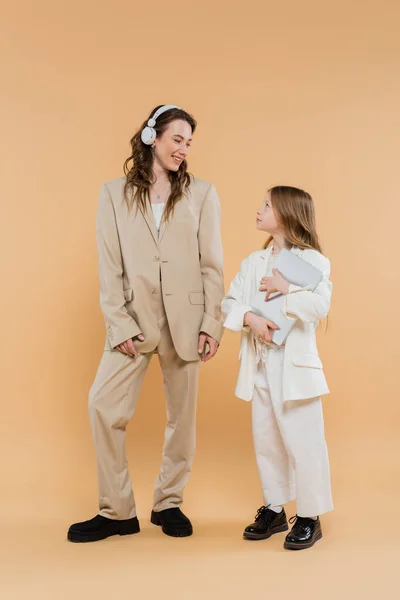 Mother and daughter in suits, woman in wireless headphones and girl with laptop on beige background, fashionable outfits, formal attire, corporate mom, modern family, online education — Stock Photo