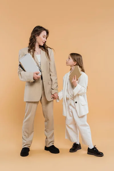 Trendy mother and daughter in suits, woman and child holding books and notebooks while standing together on beige background, fashionable outfits, formal attire, corporate mom, education concept — Stock Photo