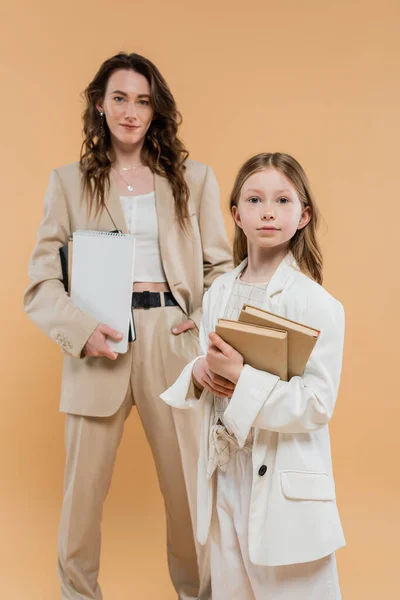 Trendy mother and daughter in suits, child holding books near blurred woman with notebooks on beige background, fashionable outfits, formal attire, corporate mom, education concept — Stock Photo