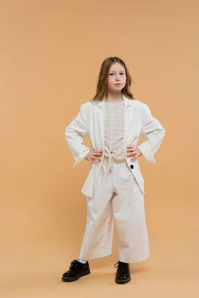 Trendy preteen girl in white suit and black shoes looking at camera while standing on beige background, fashionable outfit, formal attire, child model, trendsetter, style, posing with hands on hips — Stock Photo
