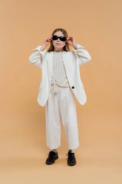 Menina pré-adolescente elegante em terno branco e sapatos pretos olhando para a câmera enquanto vestindo óculos de sol e em pé sobre fundo bege, roupa elegante, traje formal, modelo infantil, trendsetter, estilo — Fotografia de Stock
