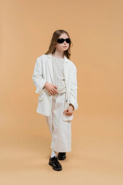 Trendy preteen girl in white suit, sunglasses and black shoes posing and standing on beige background, fashionable outfit, formal attire, child model, trendsetter, style, fashionista — Stock Photo