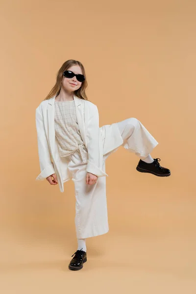 Trendy preteen girl in white suit, sunglasses and black shoes posing with raised leg and standing on beige background, fashionable outfit, formal attire, child model, trendsetter, style — Stock Photo