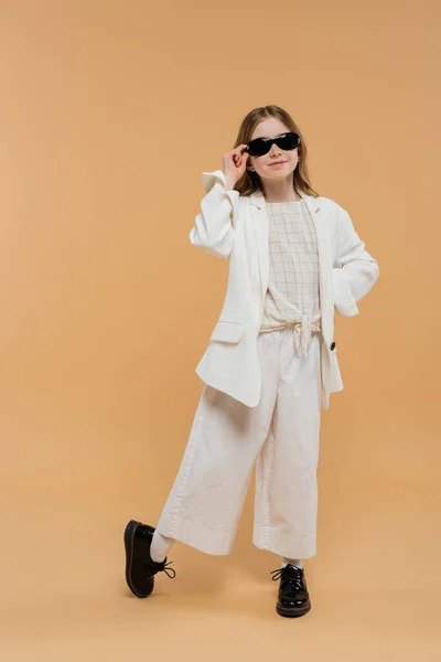 Fröhliches Mädchen im weißen Anzug, Sonnenbrille und schwarzen Schuhen posiert und steht auf beigem Hintergrund, modisches Outfit, formelle Kleidung, Kindermodel, Trendsetter, Stil, junge Eleganz — Stockfoto