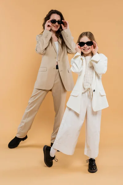 Stilvolle Mutter und Tochter mit Sonnenbrille, glückliche Geschäftsfrau und Mädchen in Anzügen, die zusammen auf beigem Hintergrund stehen, modische Outfits, formelle Kleidung, Corporate Mom, moderne Familie — Stockfoto