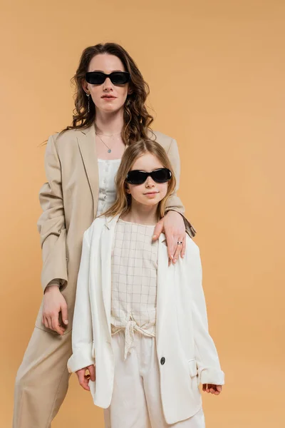 Современная семья, стильная мать и дочь в солнцезащитных очках, деловая женщина и девушка в костюмах, стоящие вместе на бежевом фоне, модные наряды, формальный наряд, корпоративная мама — стоковое фото
