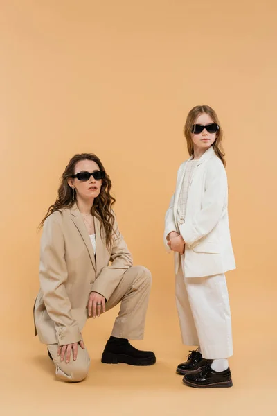 Modern family, stylish mother and daughter in suits and sunglasses, businesswoman sitting near girl on beige background, fashionable outfits, formal attire, corporate mom — Stock Photo