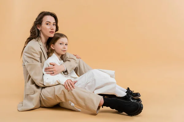 Mother child bonding concept, stylish woman in suit hugging preteen daughter and standing together in suits on beige background, corporate mom, businesswoman, motherly love — Stock Photo