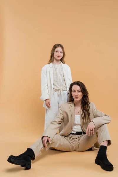 Corporate mom concept, stylish woman in suit sitting near preteen daughter and looking at camera on beige background, togetherness businesswoman, motherly love — Stock Photo