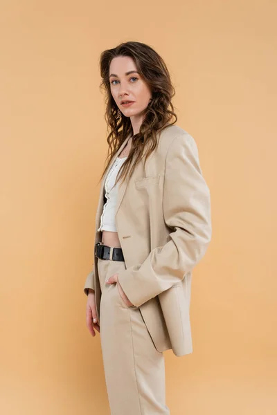 Style and fashion concept, young woman with wavy hair standing in fashionable suit while posing on beige background, formal attire, hand in pocket, looking at camera, modern elegance — Stock Photo