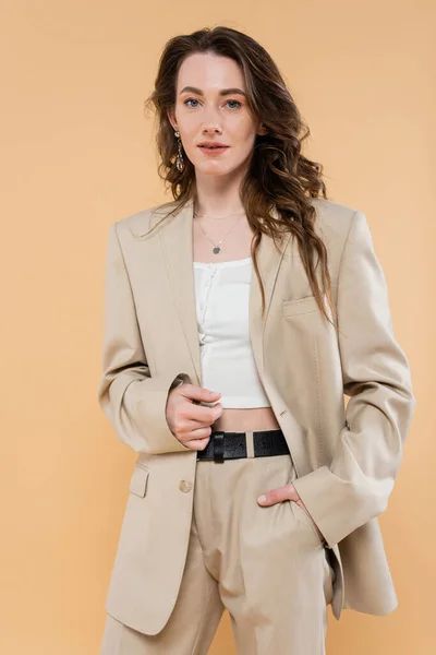Concepto de tendencia de moda, mujer joven con el pelo ondulado de pie en traje de moda y mirando a la cámara en fondo beige, mano en el bolsillo, estilo clásico, pose elegante - foto de stock