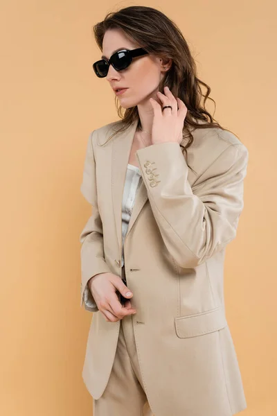 Concepto de tendencia de moda, mujer joven con el pelo ondulado de pie en traje de moda y gafas de sol sobre fondo beige, estilo clásico, elegante pose elegante, atuendo profesional, atuendo formal - foto de stock