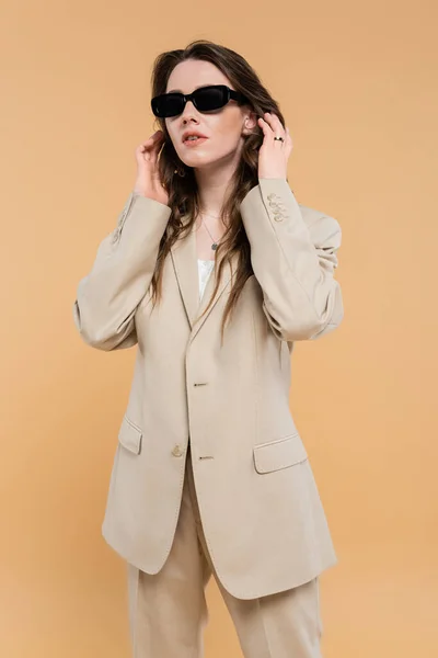 Conceito de tendência de moda, jovem mulher ajustando o cabelo ondulado e de pé em terno elegante com óculos de sol no fundo bege, estilo clássico, elegante posando, traje profissional — Fotografia de Stock