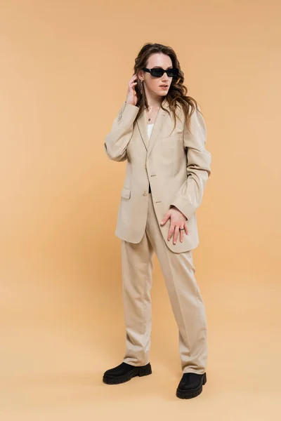 Style and fashion concept, young woman adjusting wavy hair and standing in fashionable suit with sunglasses on beige background, classic style, chic stylish posing, professional attire, full length — Stock Photo