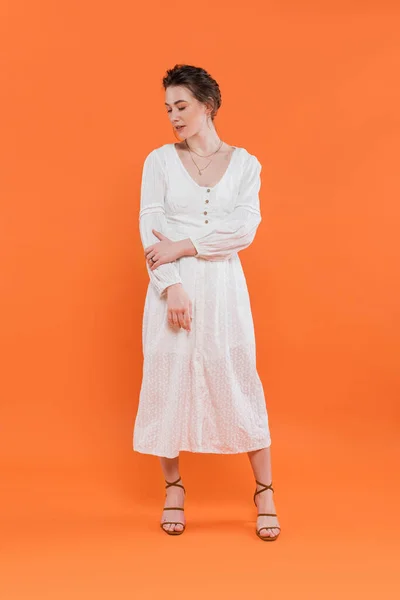 Moda de verano, mujer joven en vestido de sol blanco mirando hacia otro lado y de pie sobre fondo naranja, fondo vibrante, posando con estilo, señora de blanco, tendencia de la moda, elegancia - foto de stock