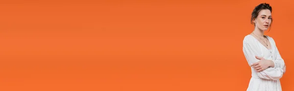 Tendances estivales, jeune femme en robe de soleil blanche regardant la caméra et debout avec les bras croisés sur fond orange, couleurs vives, pose élégante, dame en blanc, mannequin, bannière — Photo de stock